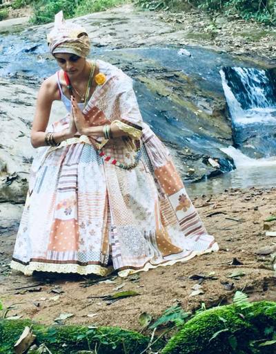 Mãe Estella com vestes tradicionais, de mãos unidas, junto a uma cachoeira